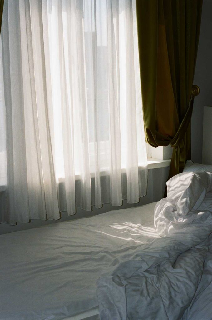 Leeres Bett mit zerknüllten Laken. Licht fällt durch die geschlossenen Vorhänge.