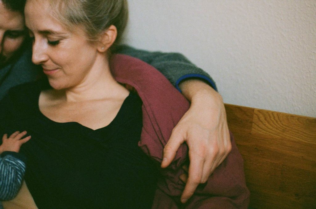 Analoges Newbornshooting in Bielefeld: Mutter im Wochenbett stillt ihr Baby. Ihr Mann legt den Arm um sie. Beide betrachten das Baby, dass nicht zu sehen ist.