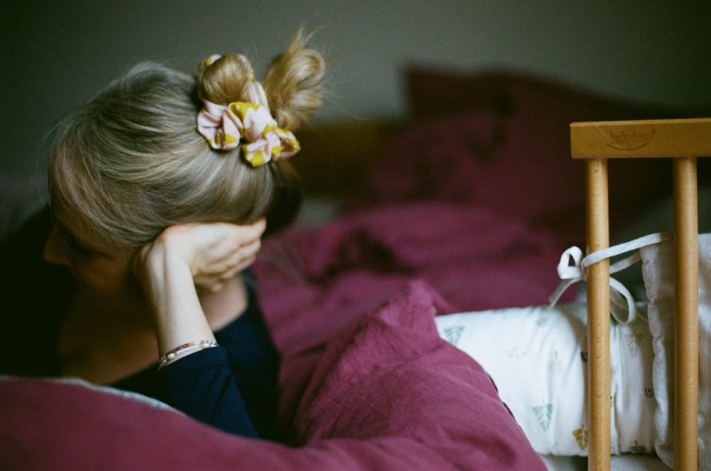 Mutter im Wochenbett liegt auf dem Bett und betrachtet ihr Baby. Sie stützt den Kopf in ihre Hände. Ihre Haare trägt sie zum Dutt.