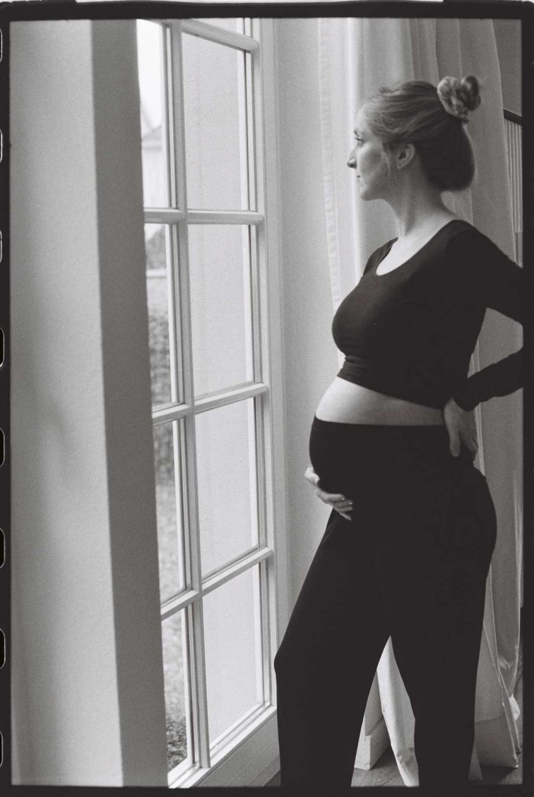 Analoges Babybauchshooting in Bielefeld: Schwangere Frau steht am Fenster und schaut nach draußen. Sie ist im Profil zu sehen, die Haare hat sie zu einem Dutt nach oben gebunden. Mit einer Hand stützt sie ihren Rücken, mit der anderen hält sie ihren Babybauch