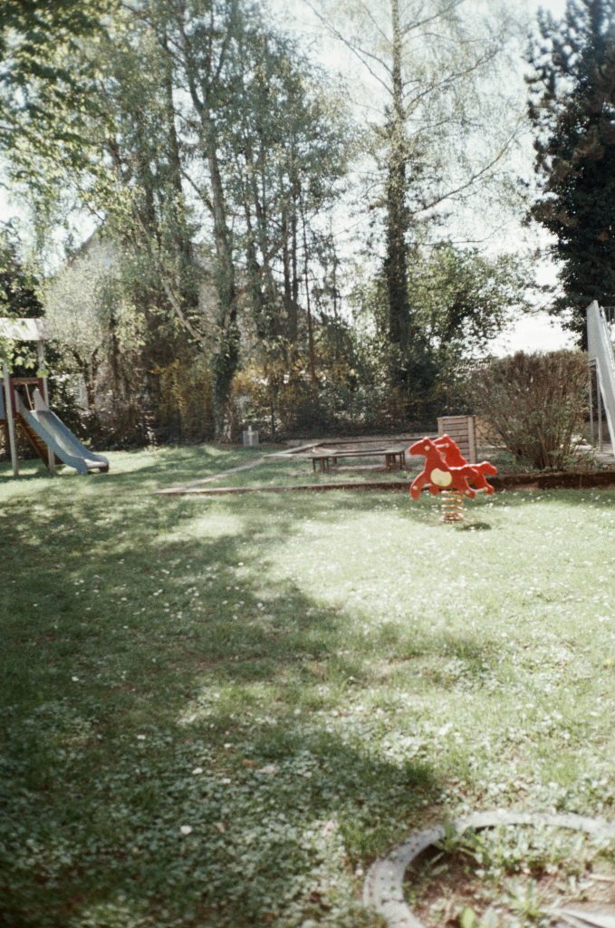 Kinderspielplatz in der Sonne. Die grüne Wiese ist mit Gänseblümchen übersät. Auf der Wiese steht eine rote Kinderwippe in Form eines Pferdes.
