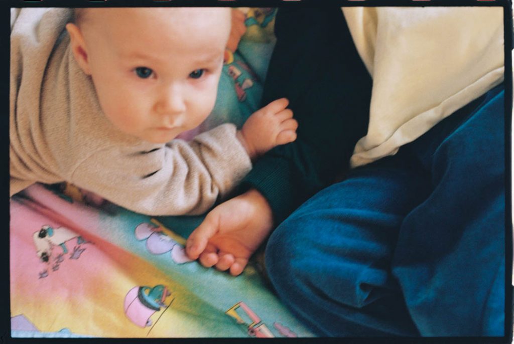 Großer Bruder mit kleiner Schwester auf dem Fußboden. Die Schwester hat ihre kleine Hand auf dem Unterarm des Bruders abgelegt und schaut in die Kamera.