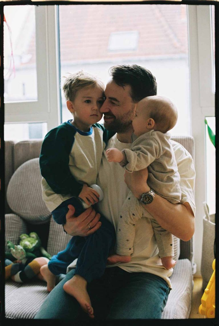 Analoges Familienshooting in Bielefeld: Vater, der mit seinen beiden Kindern auf dem Arm auf dem Boden im Kinderzimmer kniet.