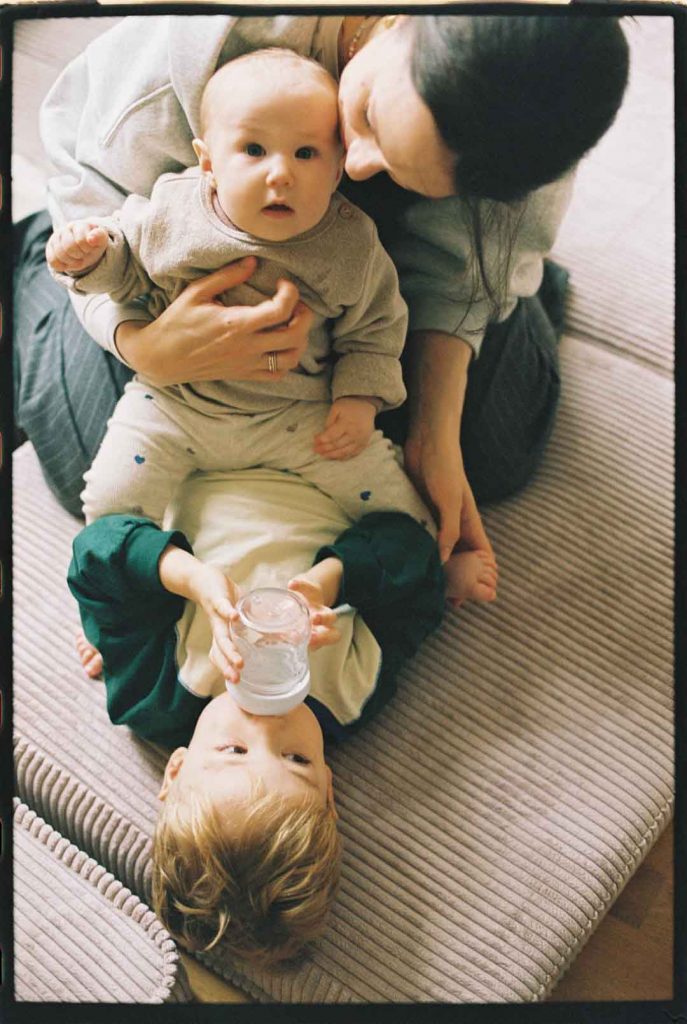 Mutter mit ihren beiden Kindern auf dem Fußboden des Kinderzimmers. Das Baby schaut in die Kamera. Das Kleinkind trinkt aus der Flasche.