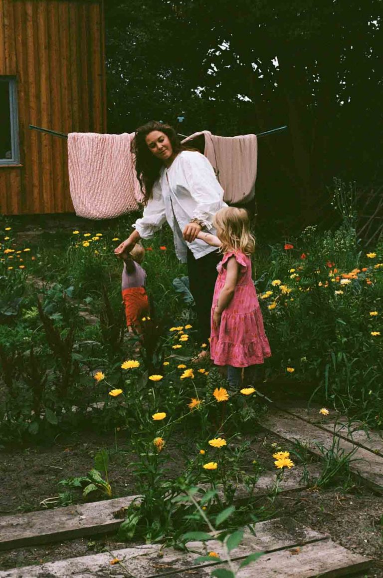 Mutter steht mit ihren zwei Kindern an der Hand im Garten. Alle haben nackte Füße und die Blumen blühen. Im Hintergrund ist eine Wäschespinne zu sehen.