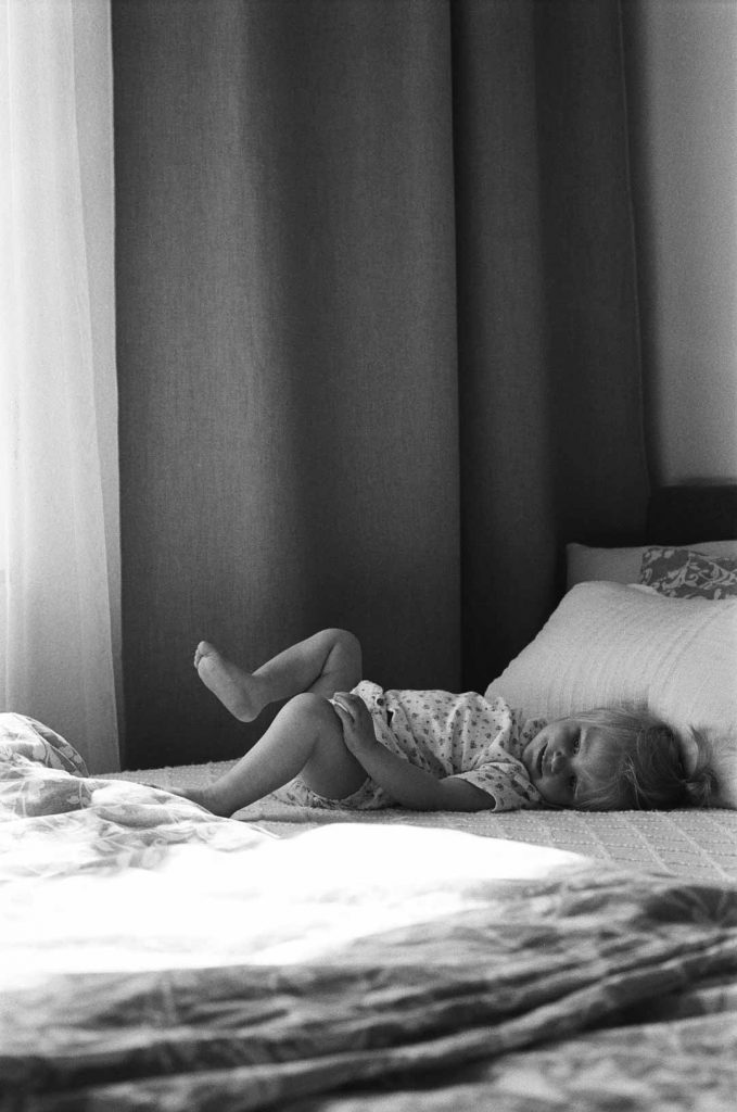 Schwarzweiß Fotografie eines kleinen Mädchen, dass auf dem Bett der Mutter liegt und lacht.