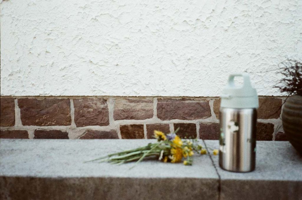 Kindertrinkflasche und selbst gepflückte Blumen, abgestellt auf einer Mauer.