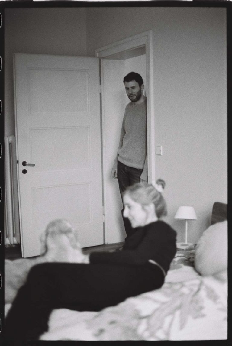 Schwangere Frau liegt auf dem Bett und und wendet sich ihrem Hund am Fußende zu. Im Türrahmen lehnt ihr Mann und beobachtet beide.