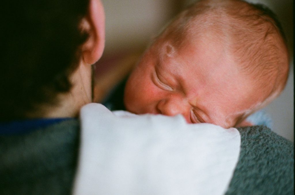 Baby im Wochenbett hat den Kopf auf der Schulter des Vaters auf einem Spucktuch abgelegt. Seine Augen sind geschlossen.
